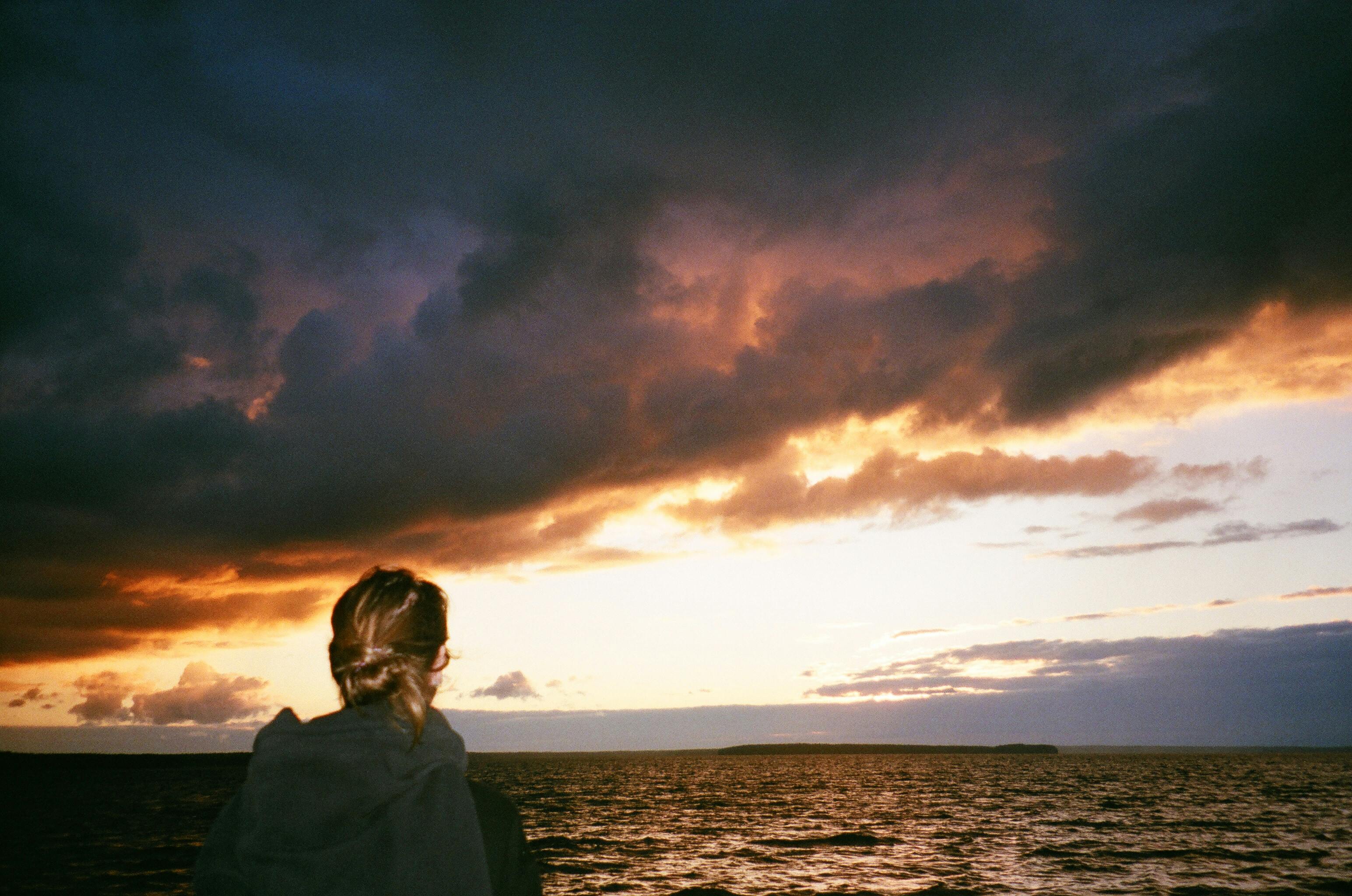 A woman gazes at a tranquil ocean sunset, surrounded by dramatic clouds. Ideal for serene and contemplative themes.