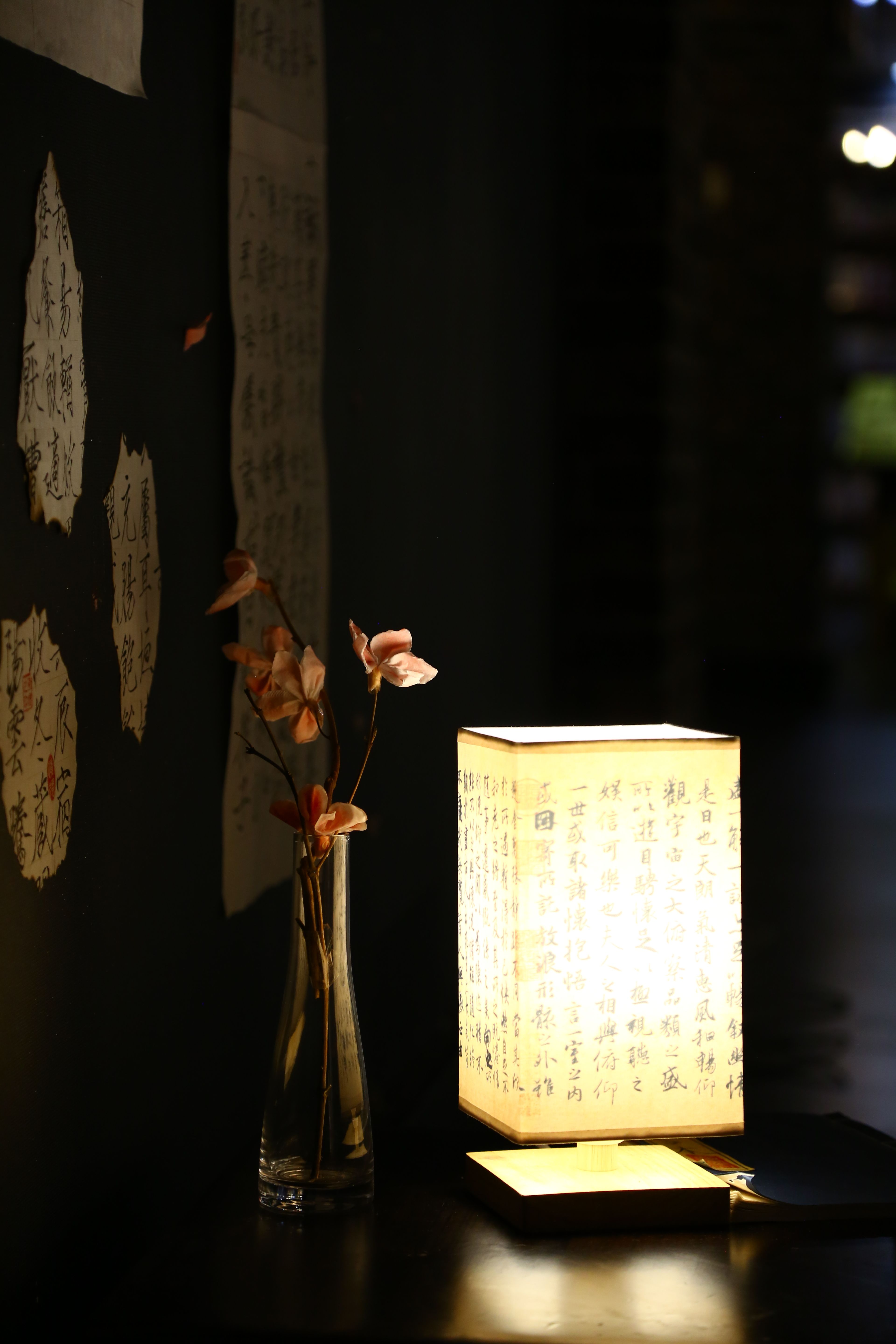 Illuminated oriental lamp with calligraphy beside a vase of flowers on a dark surface.