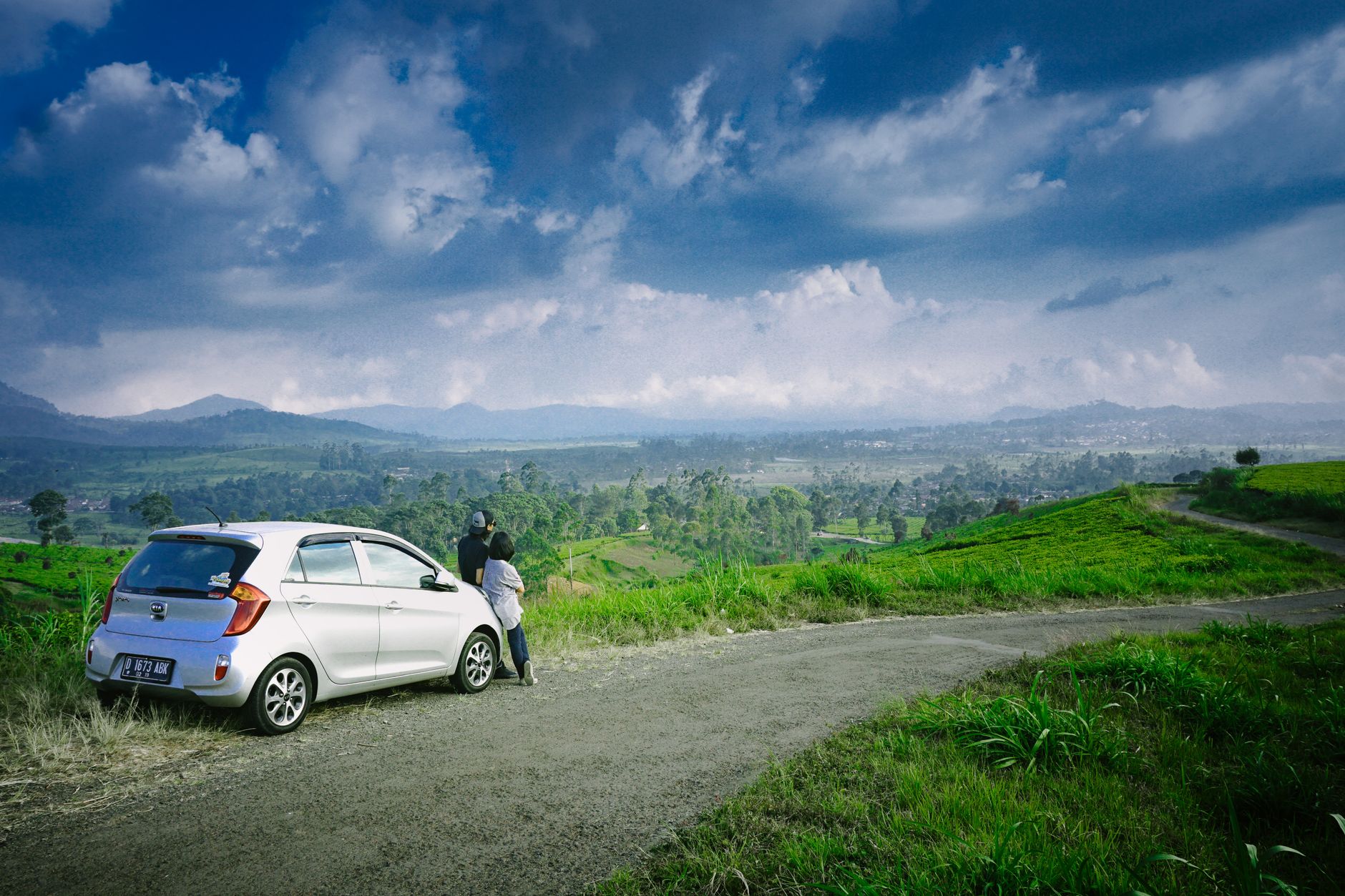 A couple stands by their car on a road overlooking lush green fields and mountains under a cloudy sky.