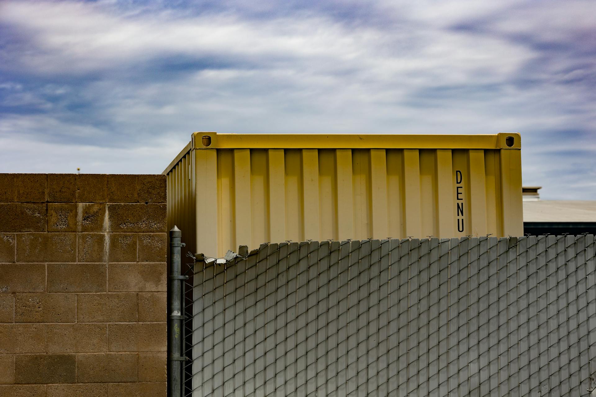 Yellow shipping container behind industrial fence under cloudy sky in urban setting.