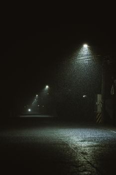 Atmospheric night scene with rain falling under street lights on an empty road, creating a dramatic ambiance.