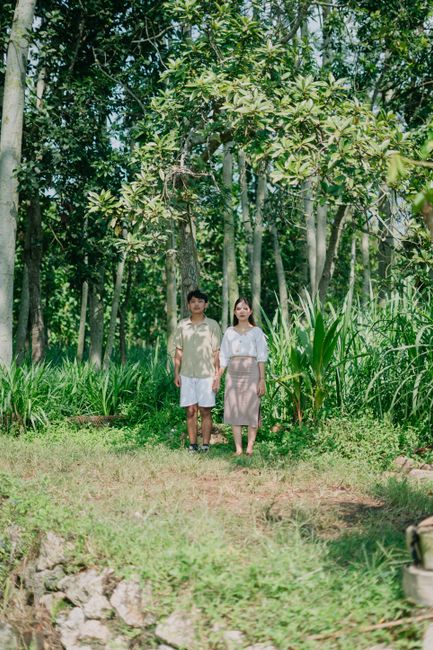 A couple standing together in a lush forest in Indonesia, under dappled sunlight.