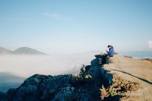 Free A hiker sitting on a cliff with a stunning view of East Java's misty mountains and blue sky. Stock Photo