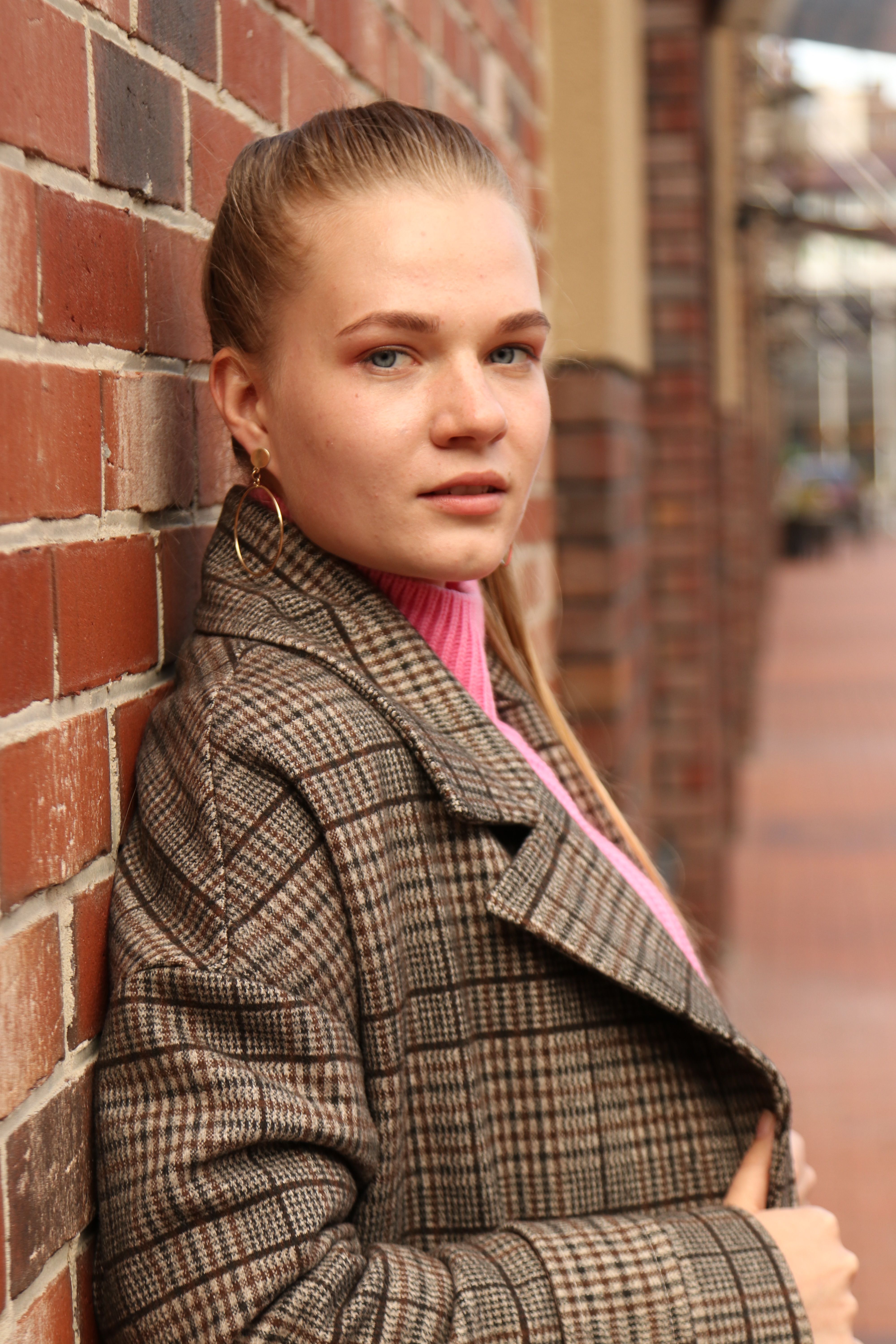 Young woman in a plaid coat leaning against a brick wall outdoors, looking at the camera.