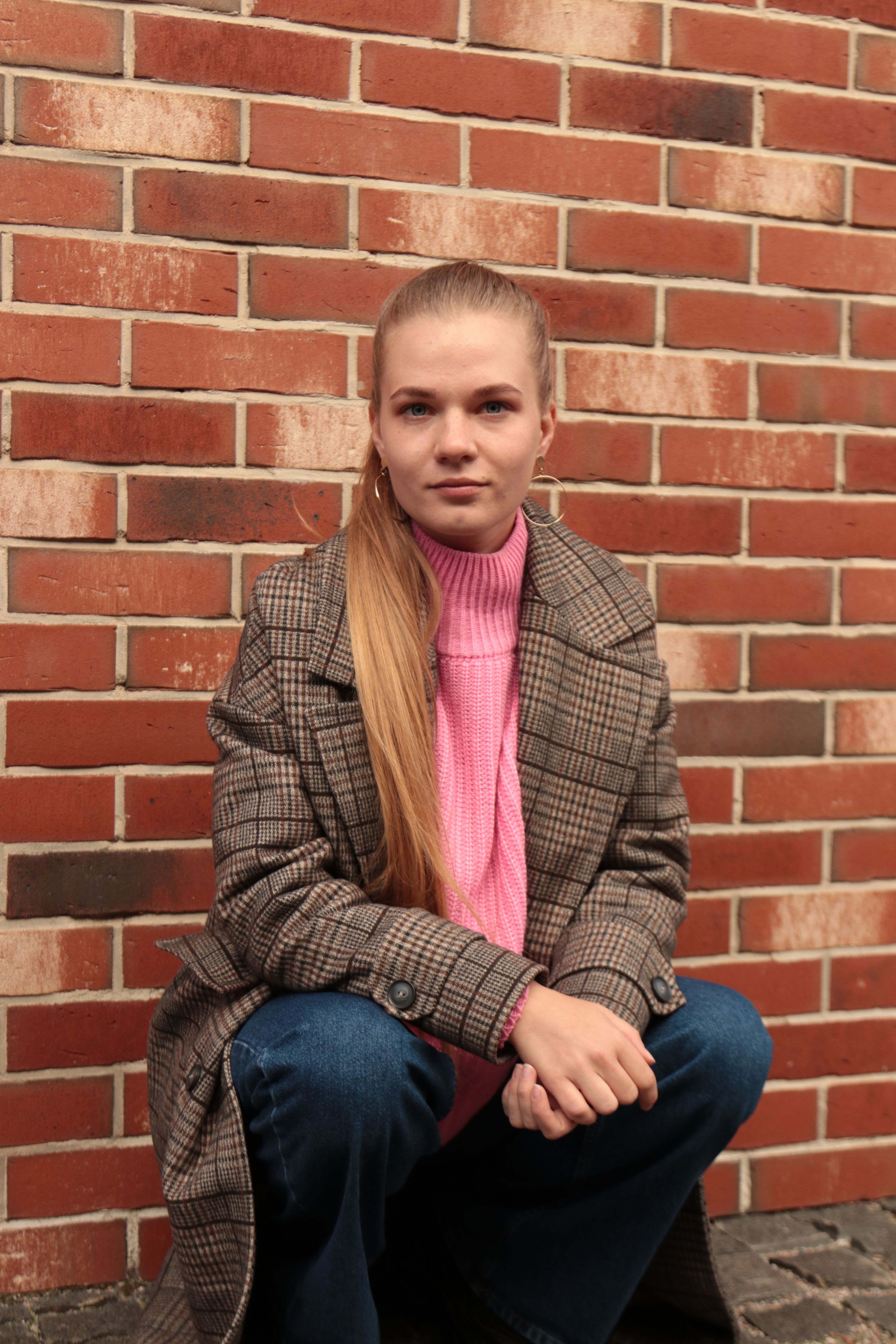 A young woman in a plaid coat, denim pants, and pink sweater crouches against a brick wall, showcasing her fashion style.