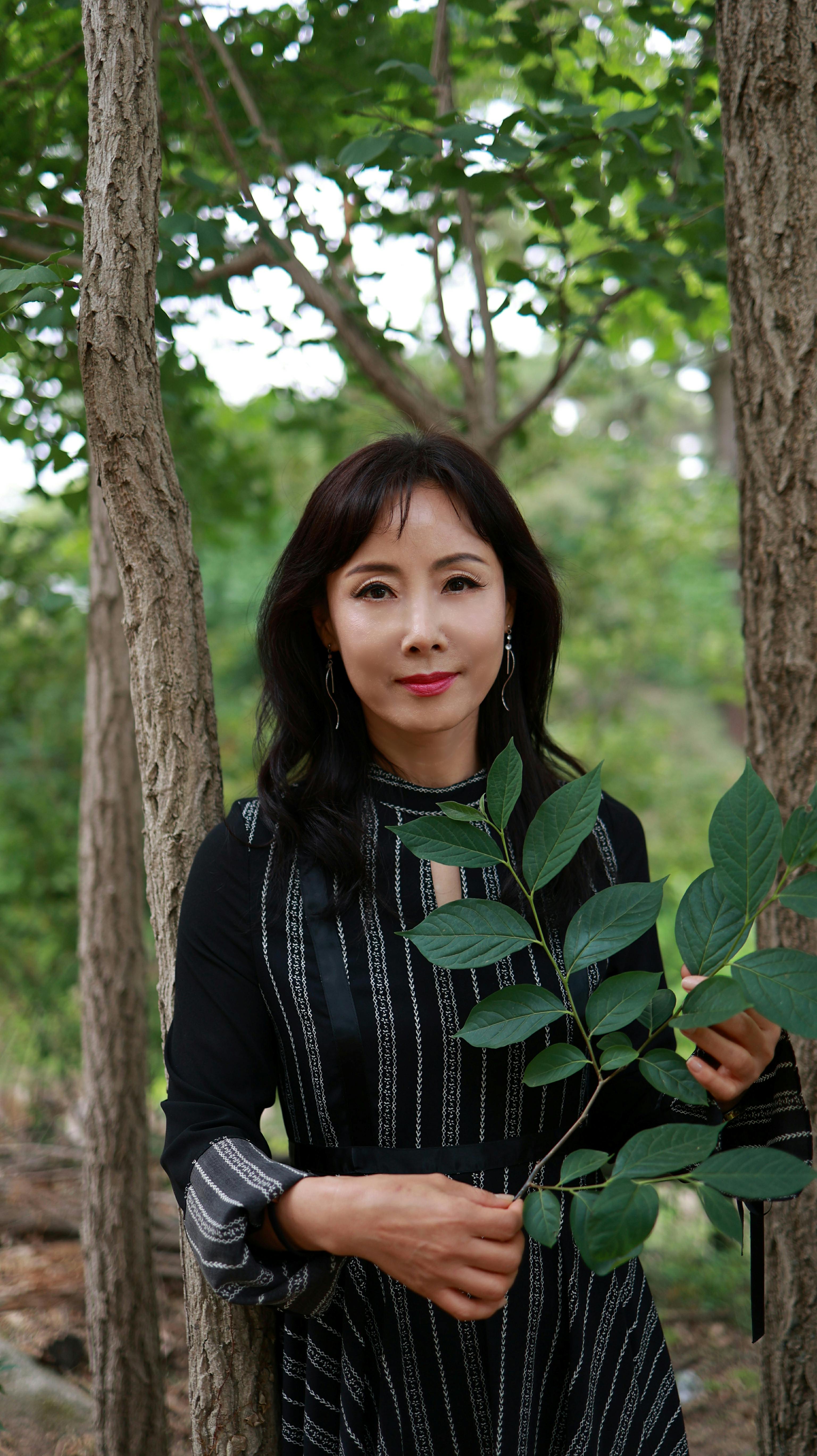 A portrait of an Asian woman in a black dress holding leaves amid a serene forest scene.