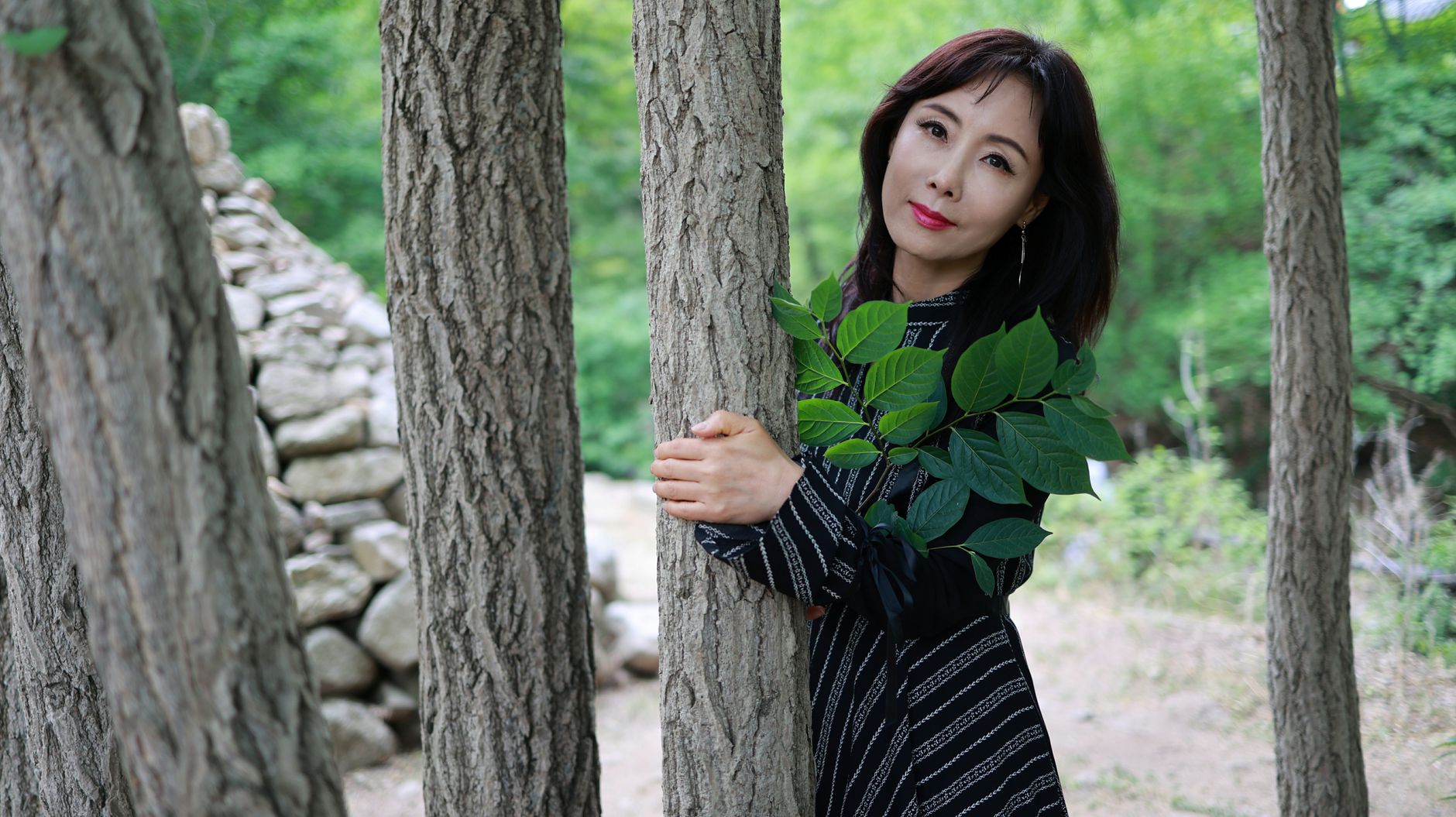 Asian woman in a black dress holding leaves in a serene forest setting, evoking harmony with nature.