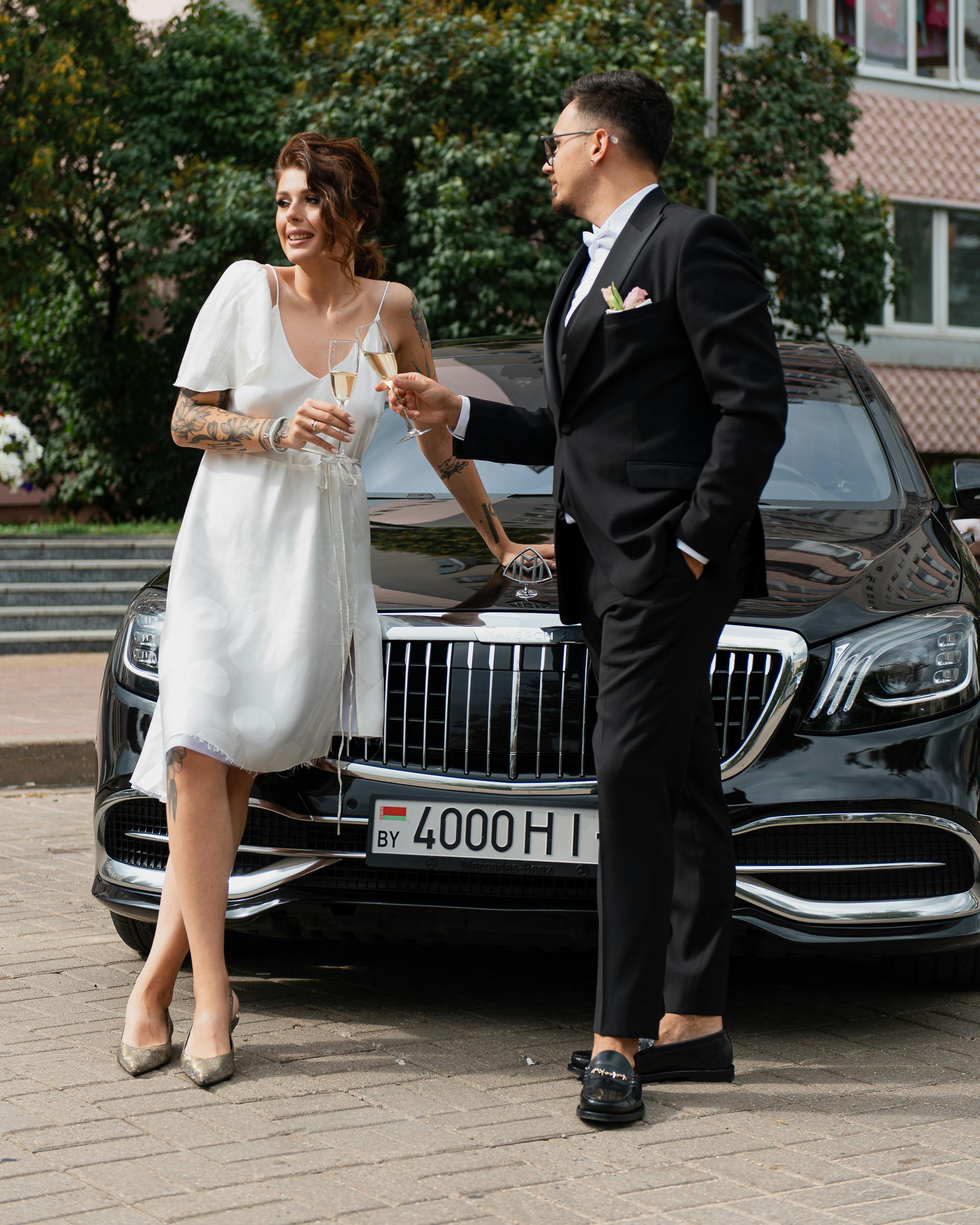 woman in white dress and man in black suit standing beside the black luxury car while drinking a glass of champagne