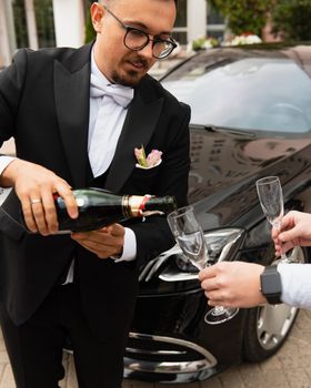 A man in formal attire pours champagne into glasses by a luxury car, conveying elegance and celebration.