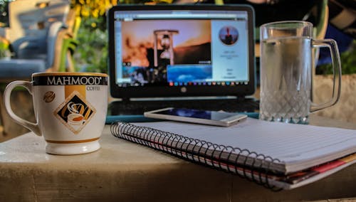 免费 陶瓷杯和玻璃啤酒杯之间的螺旋笔记本上的白色智能手机附近黑色电脑显示器显示小时玻璃 素材图片