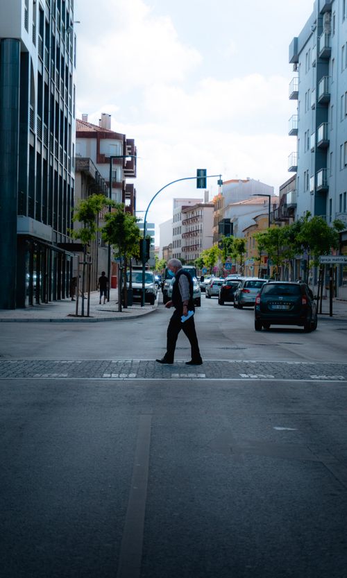 Free Elderly man crosses a city street in Portugal, wearing a mask, amidst traffic and buildings. Stock Photo