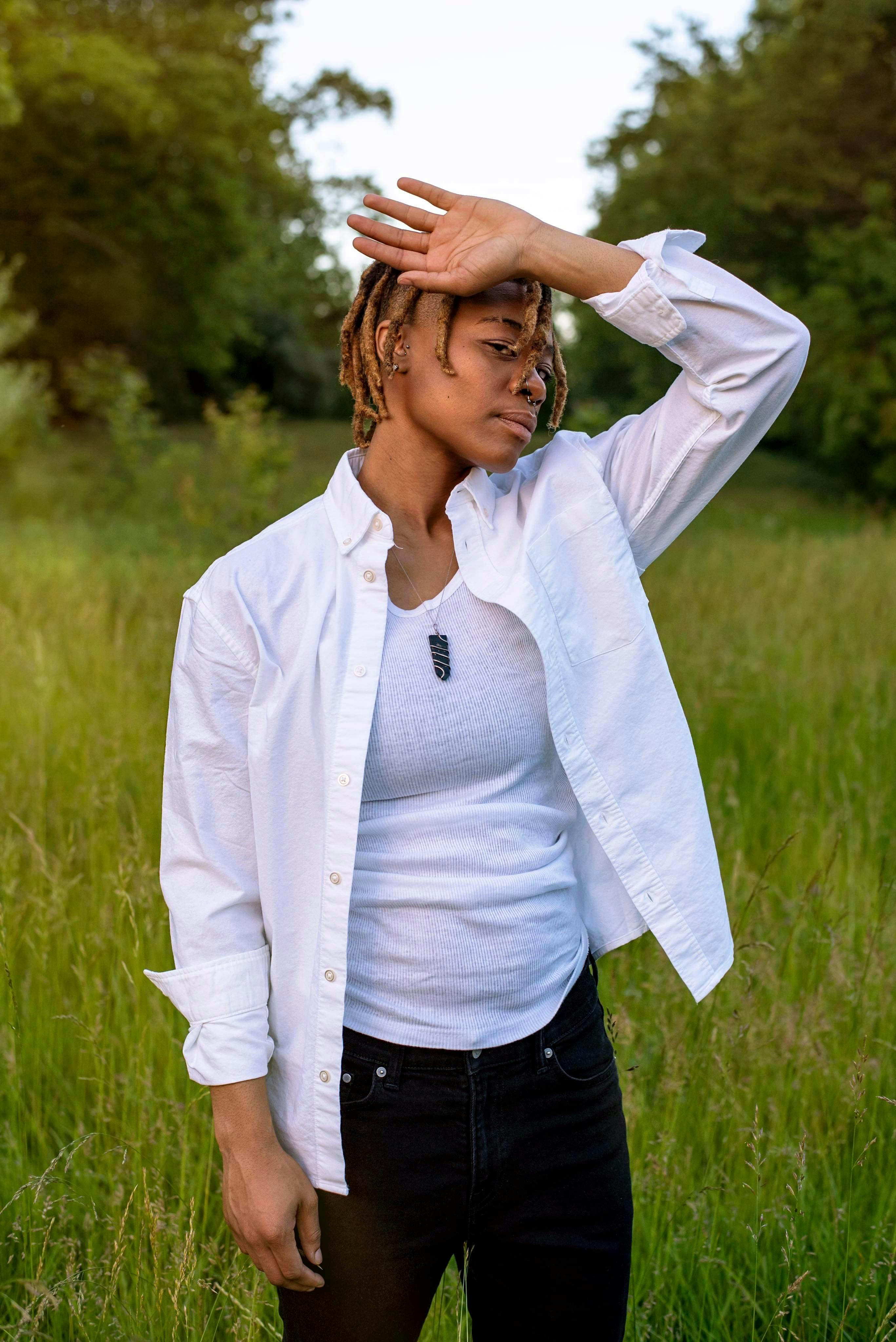 A woman in a white shirt poses under sunlight in a grassy field, expressing relaxation and style.