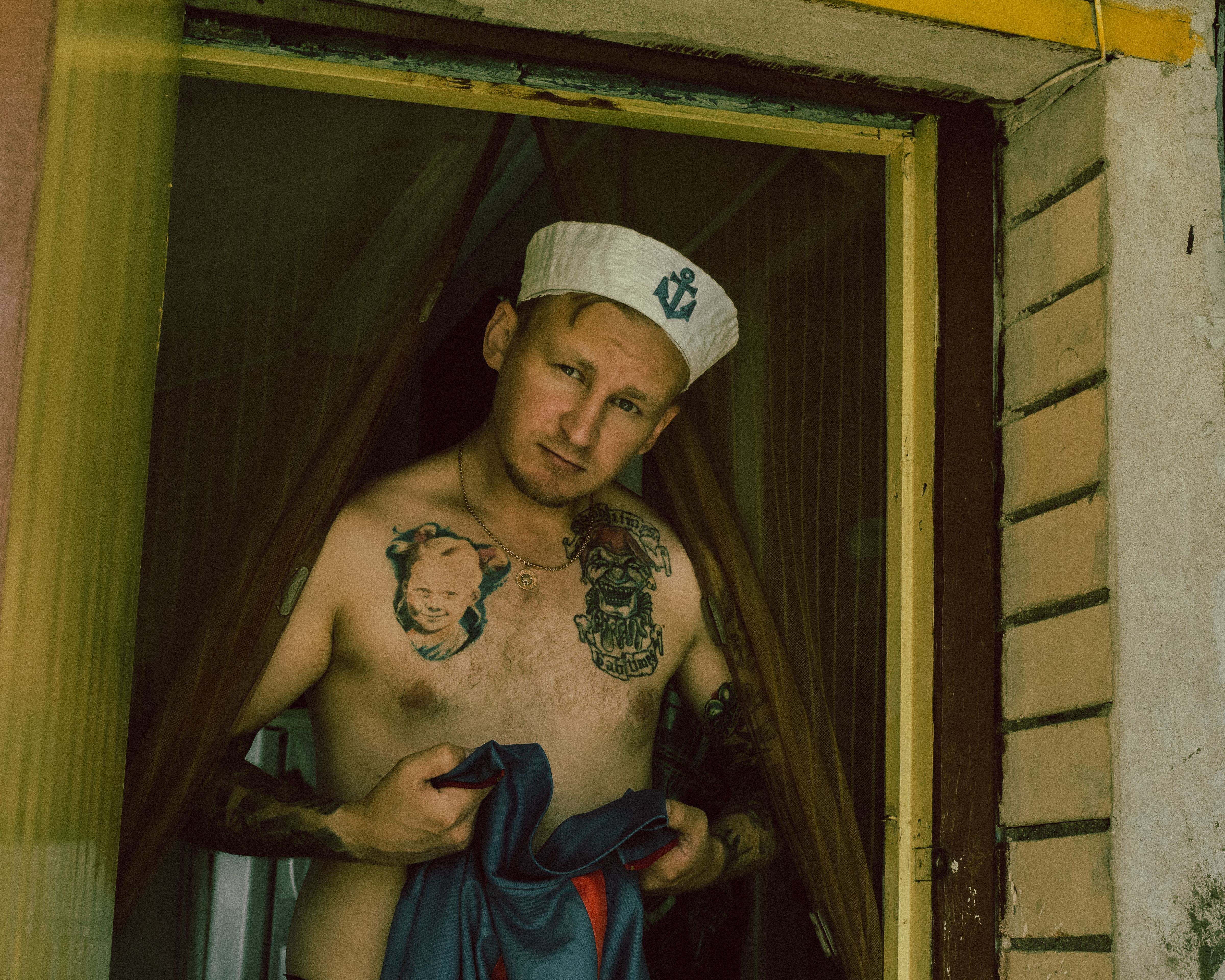 Tattooed man in sailor cap standing shirtless at a doorway with thoughtful expression.