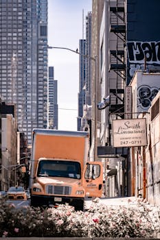 Orange delivery truck in Manhattan's urban cityscape with towering skyscrapers.