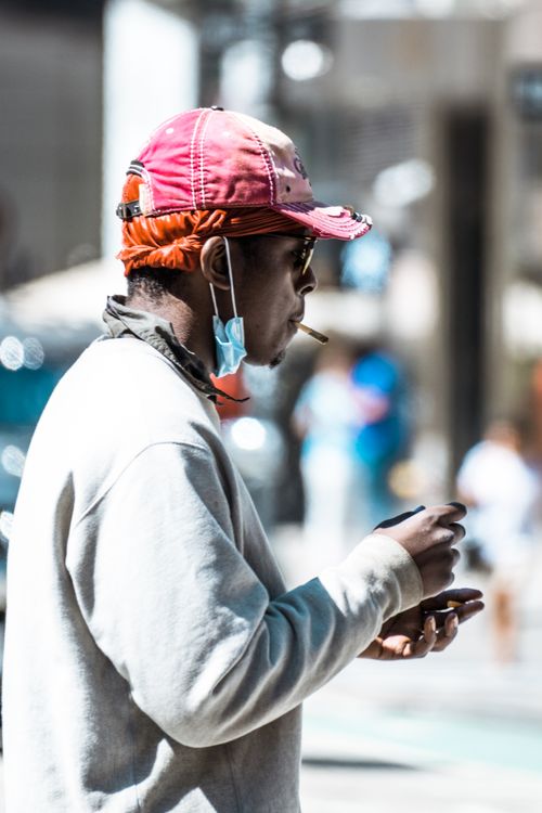 Free Side view portrait of a man smoking on an urban street, wearing a pink hat and casual attire, with a mask under his chin. Stock Photo