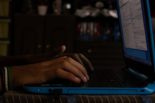 Hands typing on a blue laptop indoors, showcasing focus on work or study in a dimly lit room.