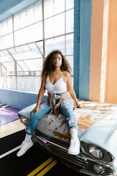 Young woman with curly hair sitting on a vintage car hood in a bright indoor setting.