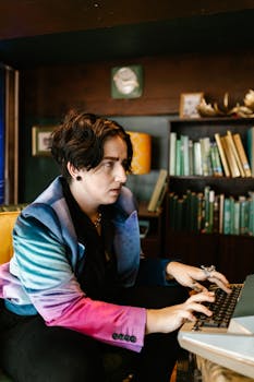 A woman in a colorful jacket types on a keyboard in a cozy room with bookshelves.