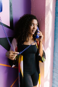 A fashionable woman with curly hair in colorful attire holds a retro blue phone indoors.