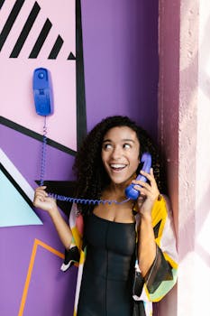 A cheerful woman with curly hair talks on a blue corded phone against a vibrant geometric wall.