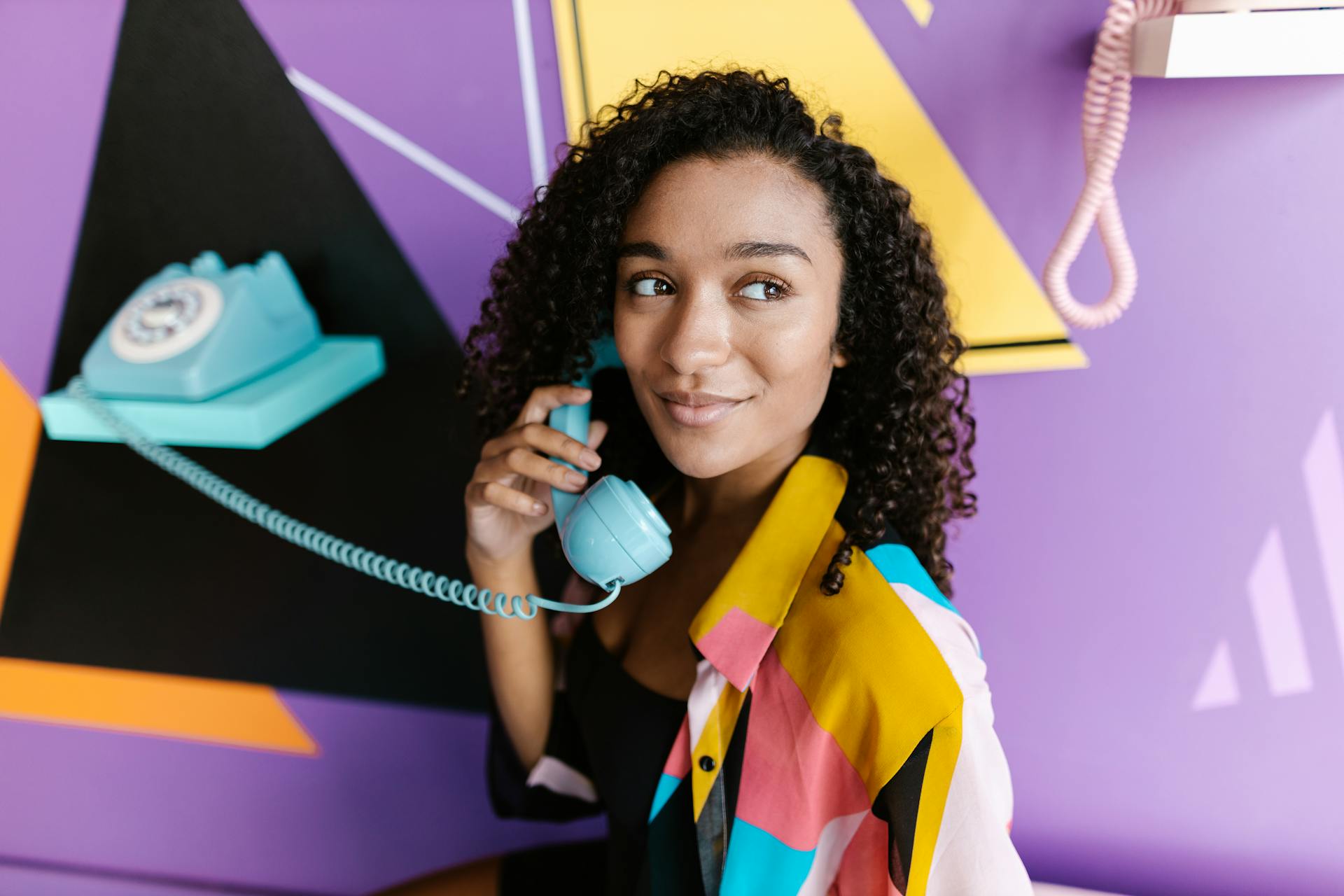 A woman with curly hair smiles while using a vintage blue telephone against a colorful backdrop.