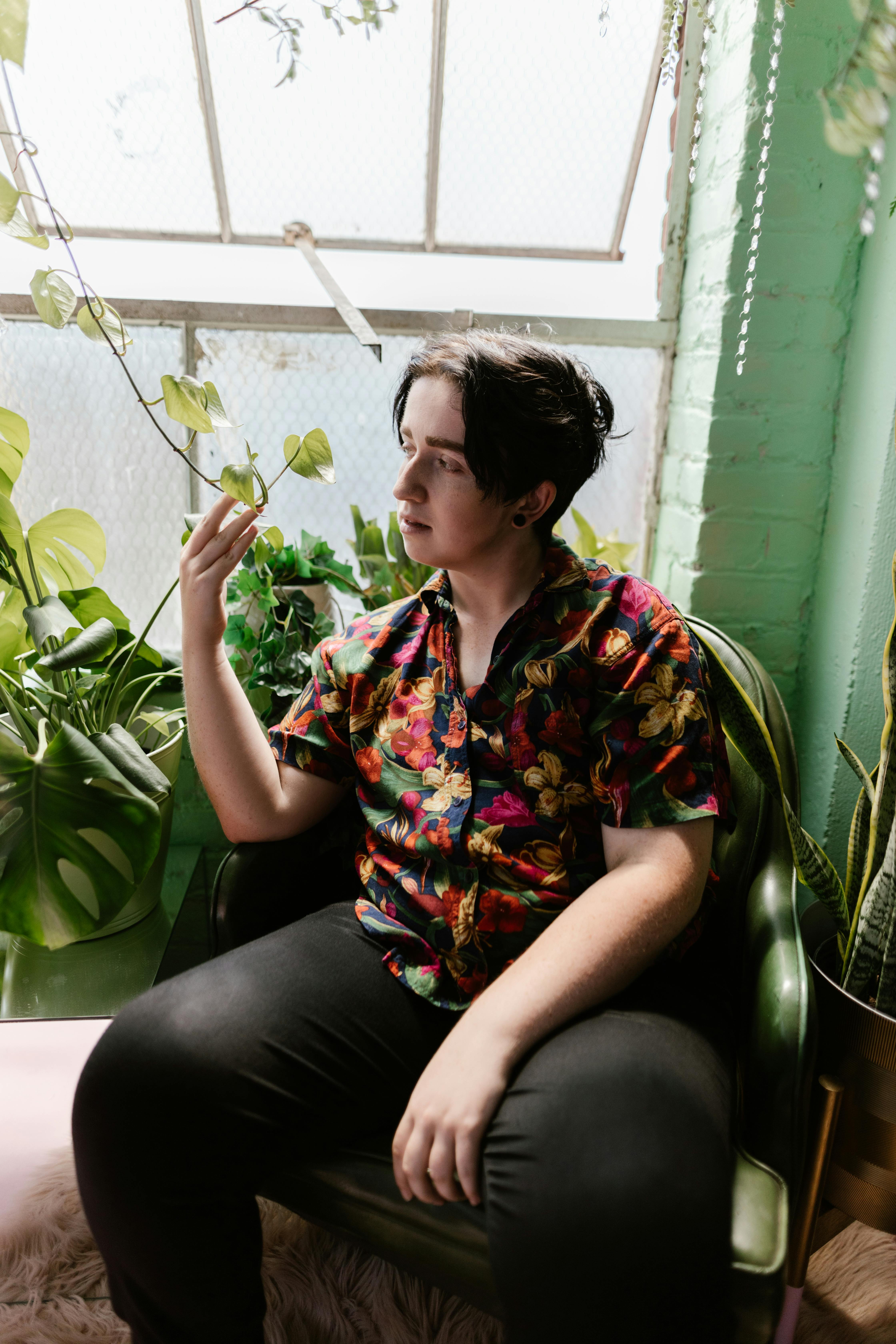 Free A young adult in a colorful shirt sits among lush indoor plants, gazing thoughtfully. Stock Photo
