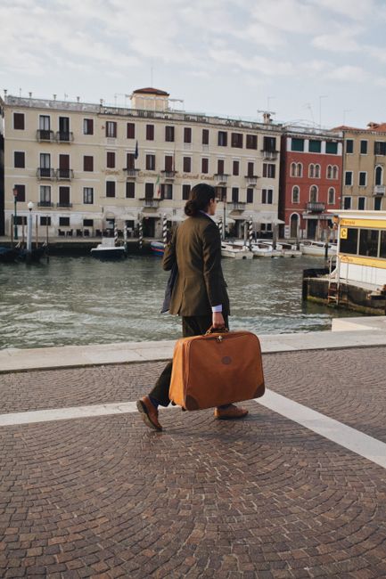 A fashionable traveler carries a suitcase along a canal in Venice, Italy, showcasing the city's iconic architecture.