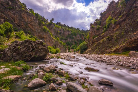 A tranquil stream flows through a green valley with rocky cliffs and cloudy skies.