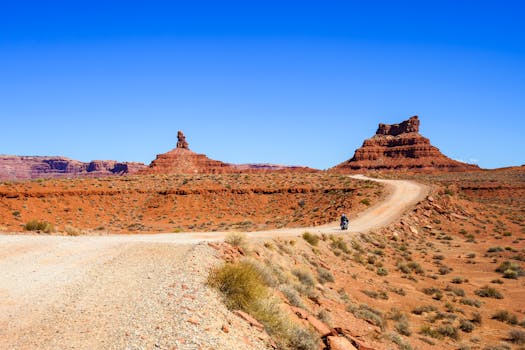 A lone motorcyclist rides along a winding dirt road in Monument Valley under a clear blue sky.