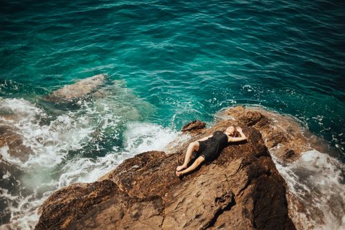 Free A woman in a black dress lying on rocky shores with the ocean waves in Karaburun, Turkey. Stock Photo