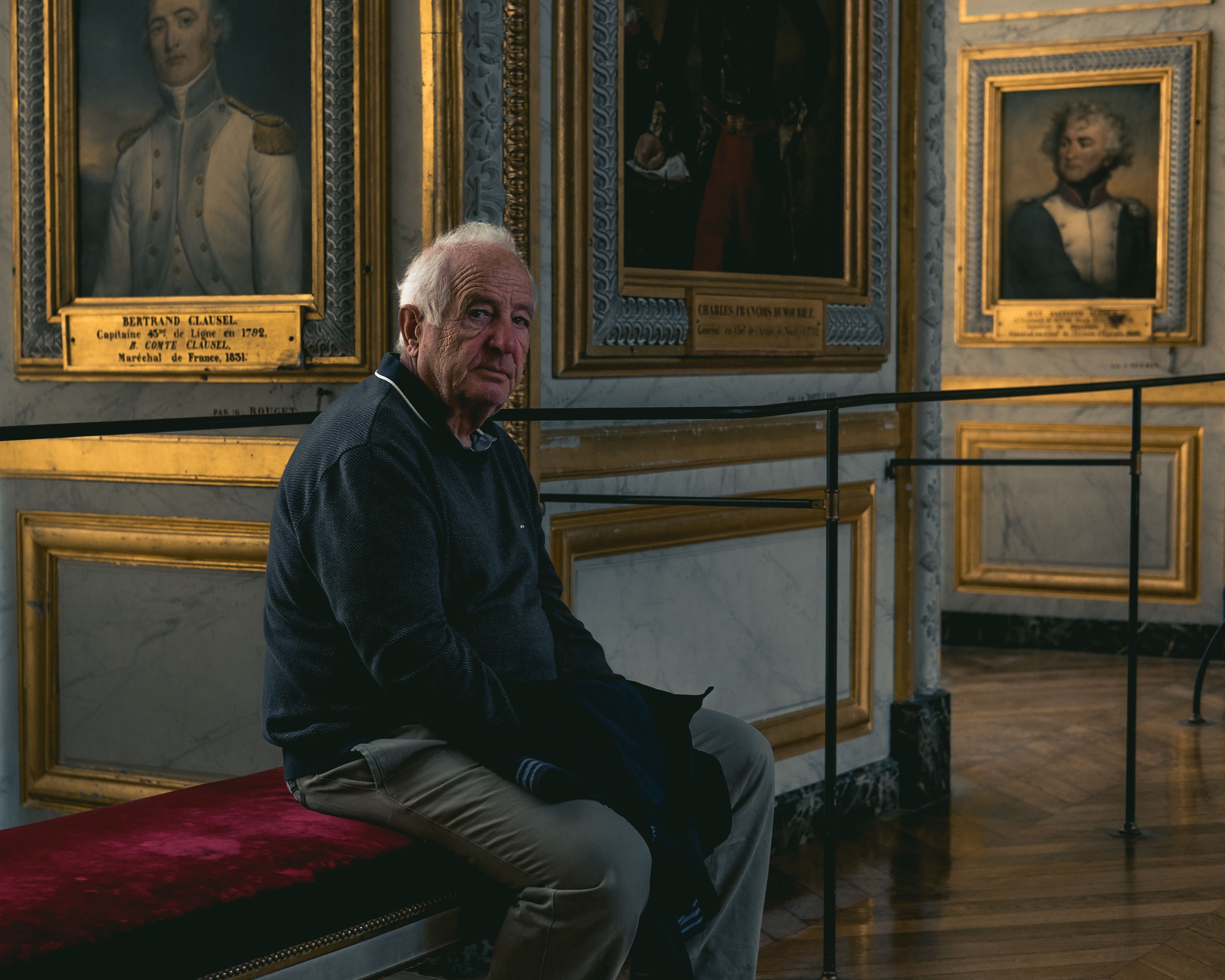 Elderly man seated on a bench in a Versailles art gallery, surrounded by historical paintings.