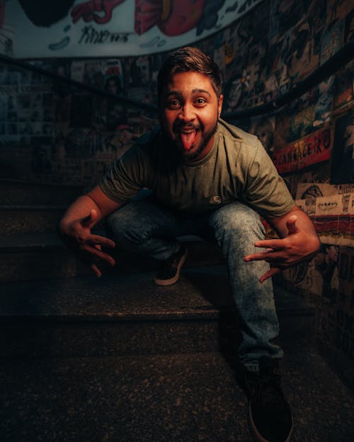 A Man in Denim Jeans Sitting on Concrete Stairs while Posing with His Tongue Out