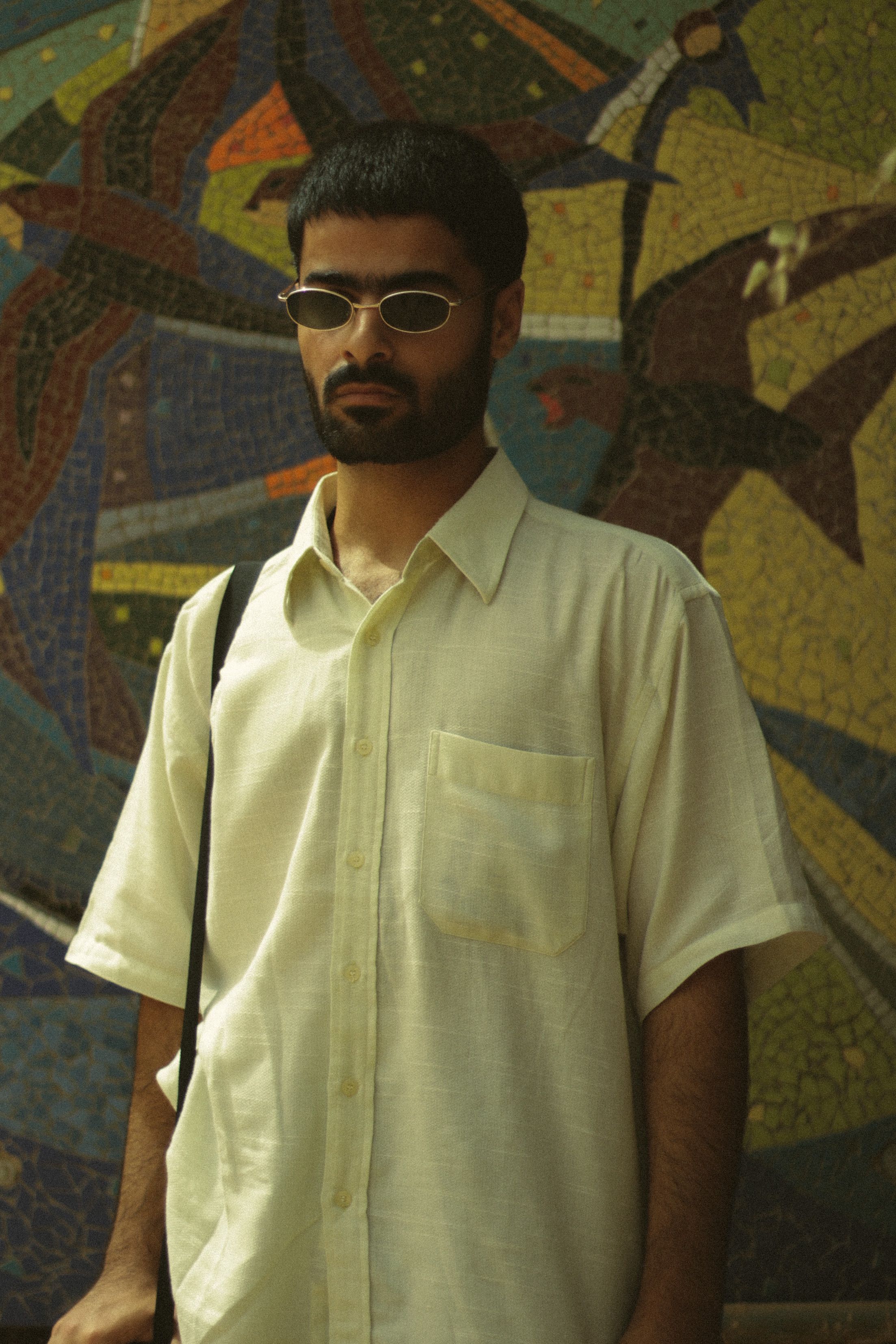 A stylish man in sunglasses stands before a colorful mosaic wall.