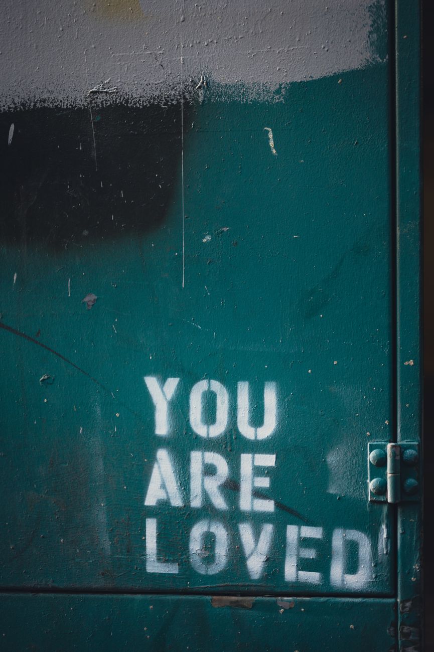 Close-up of a green painted wall featuring 'You Are Loved' text in an urban setting.