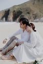 Man in White Dress Shirt and Woman in White Dress Sitting on White Sand