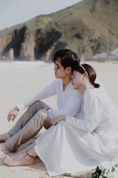 A bride and groom sitting lovingly on a beach, enjoying their wedding day.