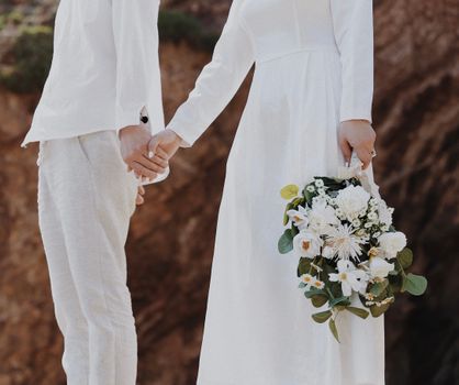 Bride and groom holding hands with white floral bouquet outdoors on wedding day.