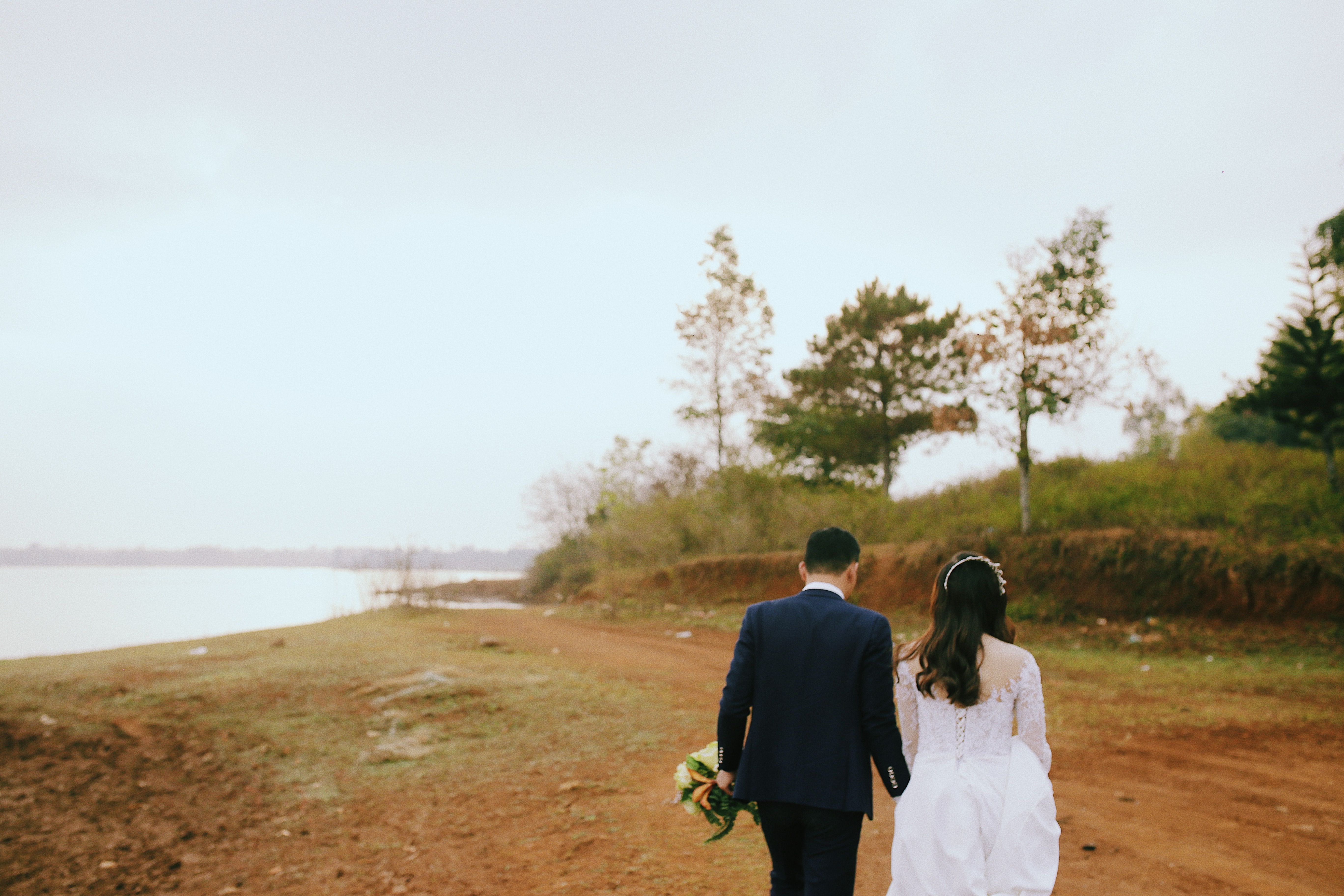 Bride and groom walking along a scenic lake shore, hand in hand, in wedding attire.