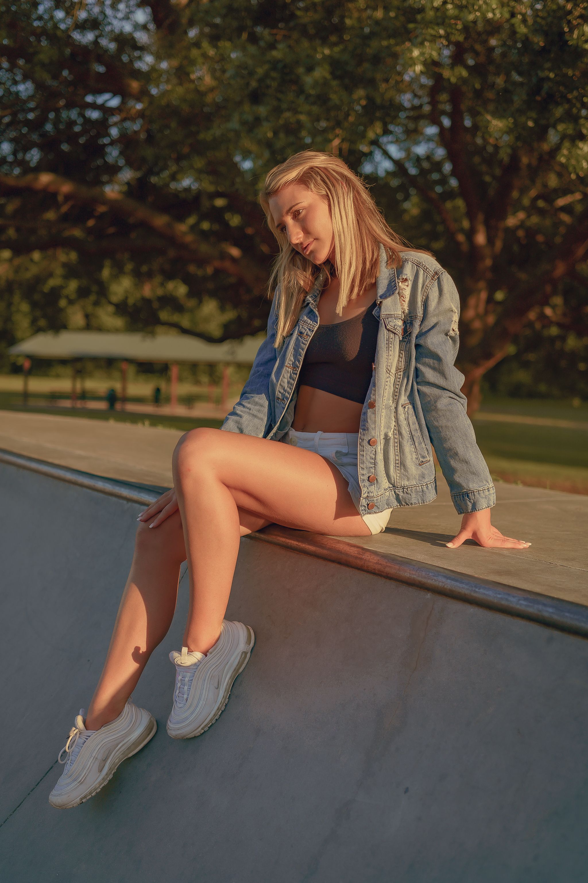 A young woman in casual summer attire relaxes by the edge of a skate park under a warm afternoon sun.