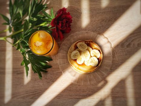 Top view of banana pancakes and orange juice with red flower, creating a warm breakfast vibe.