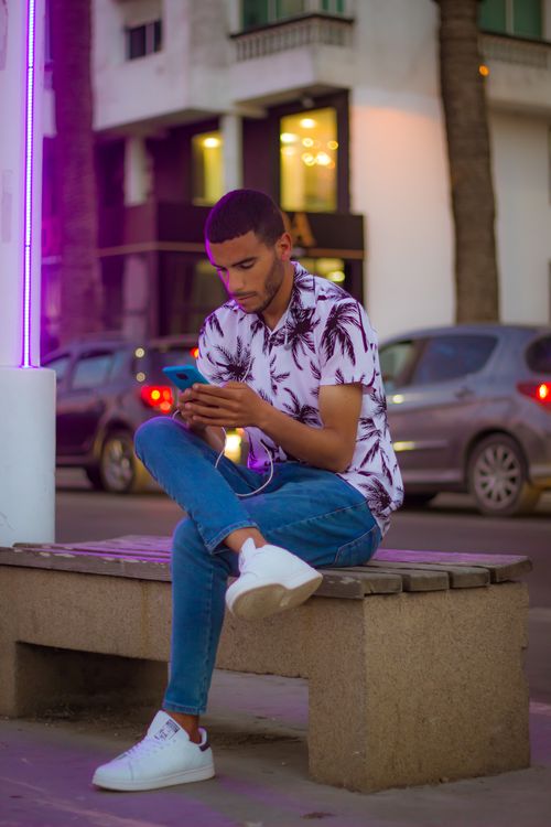 Free Young man in casual wear texting on a smartphone while sitting on a bench in urban El Jadida. Stock Photo