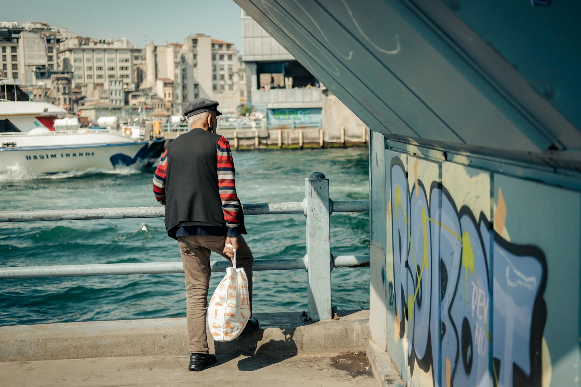 Elderly man stands by a busy city waterfront, overlooking a harbor and graffiti.