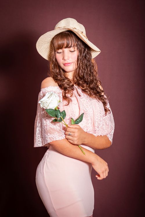 Free Charming woman in pink dress with straw hat holding a white rose against maroon background. Stock Photo