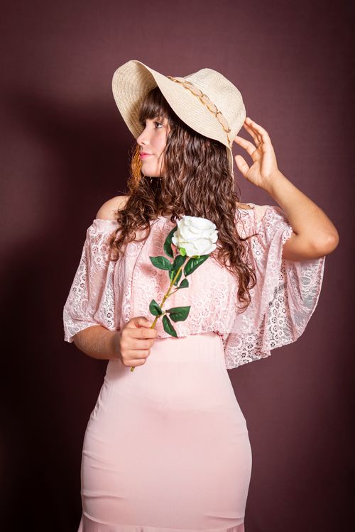 Free Elegant portrait of a woman holding a white rose, wearing a pink lace dress and hat, set against a brown backdrop. Stock Photo