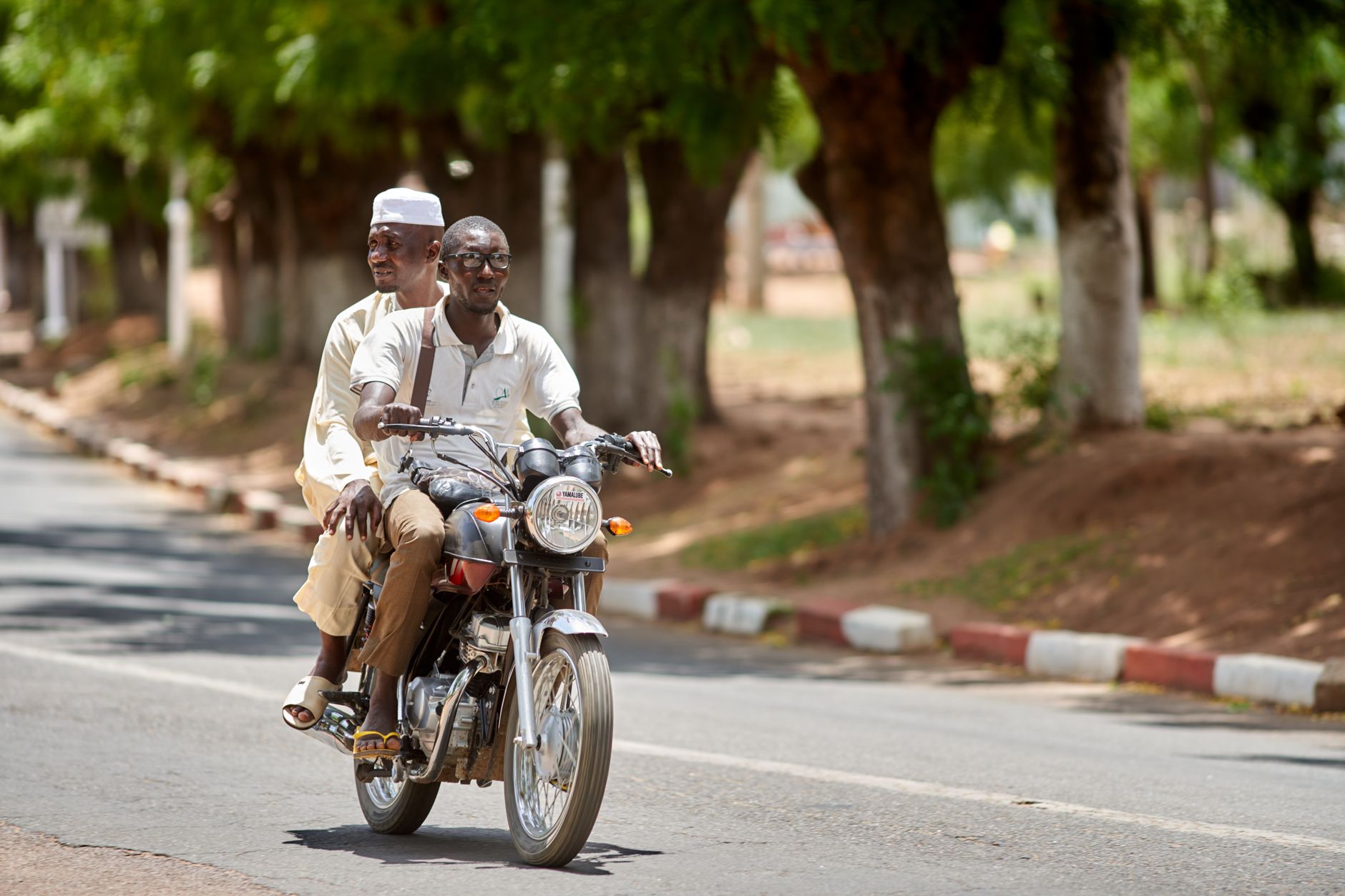 Two African men riding a motorcycle on an urban road lined with trees during the day.