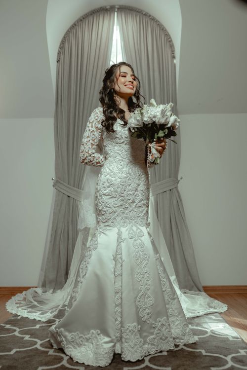 Free Young bride in a lace wedding dress holding a bouquet, smiling indoors. Stock Photo