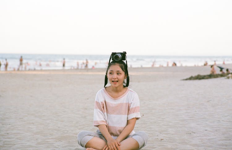 A Girl Sitting On The Beach Sand With A Camera Over Her Head