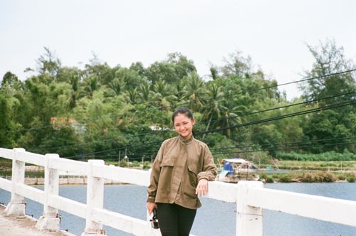 Free A woman in a brown jacket smiles on a white bridge with lush greenery and a river in the background. Stock Photo