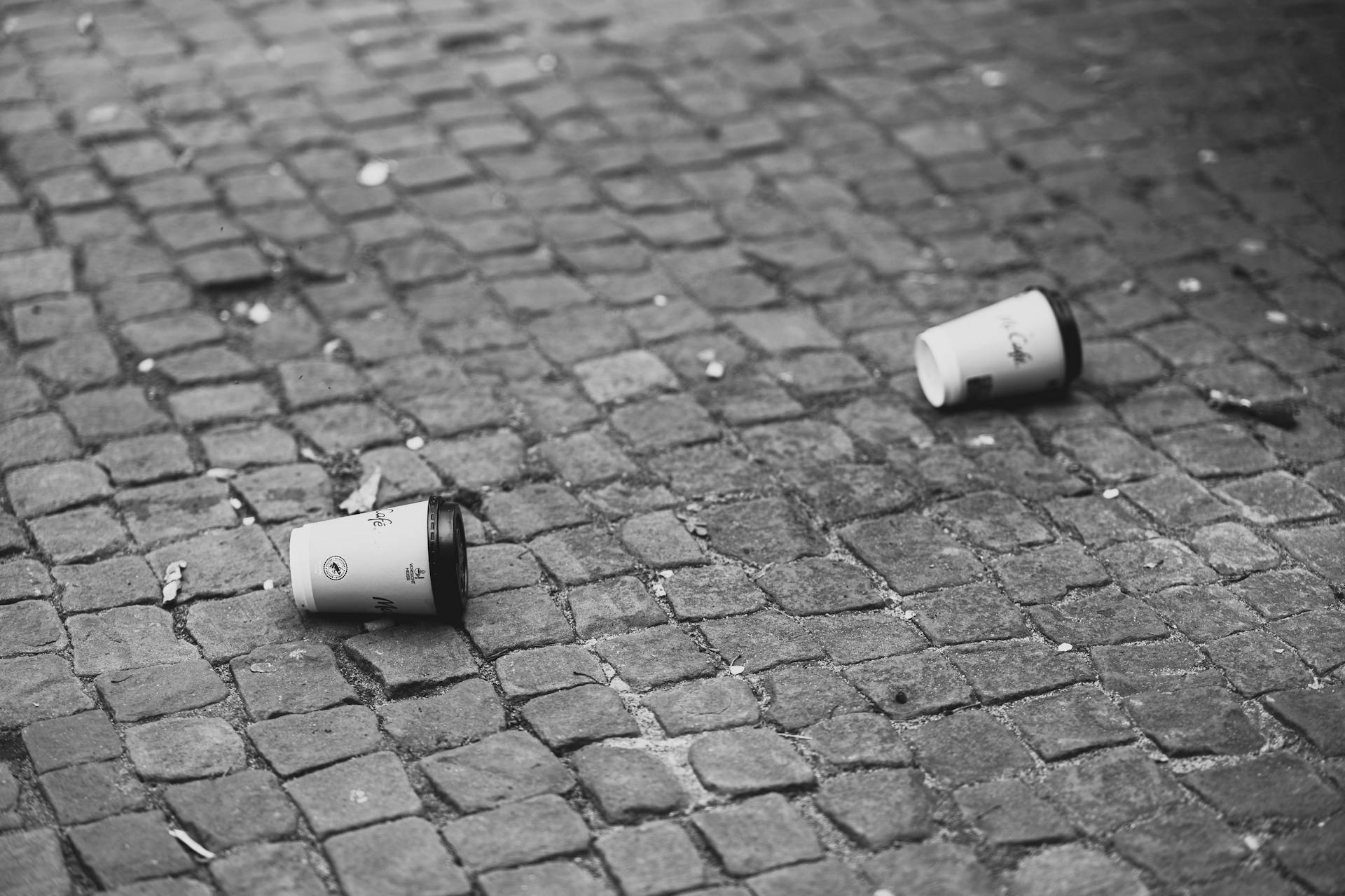 Monochrome photo capturing disposable cups littered on a cobblestone pavement.