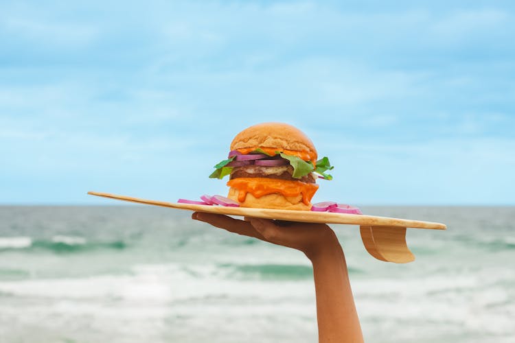 Hand Holding Board With Burger On Sea Background